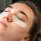 Una mujer se relaja en una cama, con parches en los ojos mientras se prepara para las extensiones de pestañas, mostrando un momento de cuidado personal.