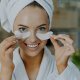 Una mujer en bata de baño sostiene una mascarilla facial y muestra sus parches oculares de hidrogel para una refrescante rutina de cuidado de la piel.